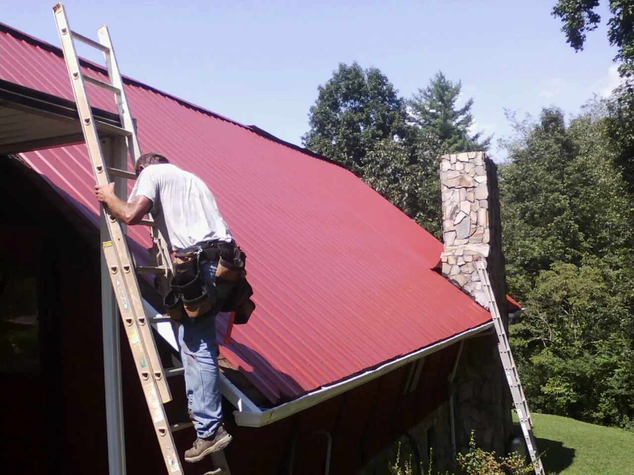 Roofer on a ladder beside a red metal roof and stone chimney — Soffit & Fascia Repair by Roofing Brothers in Donahue